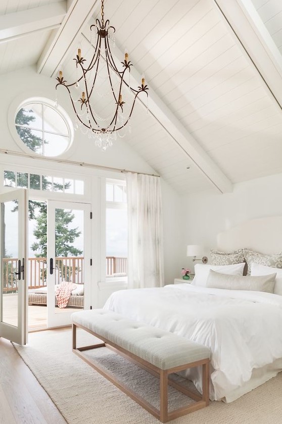 a beautiful white attic bedroom with a white bed, a grey bench, printed textiles and a blacony with a view