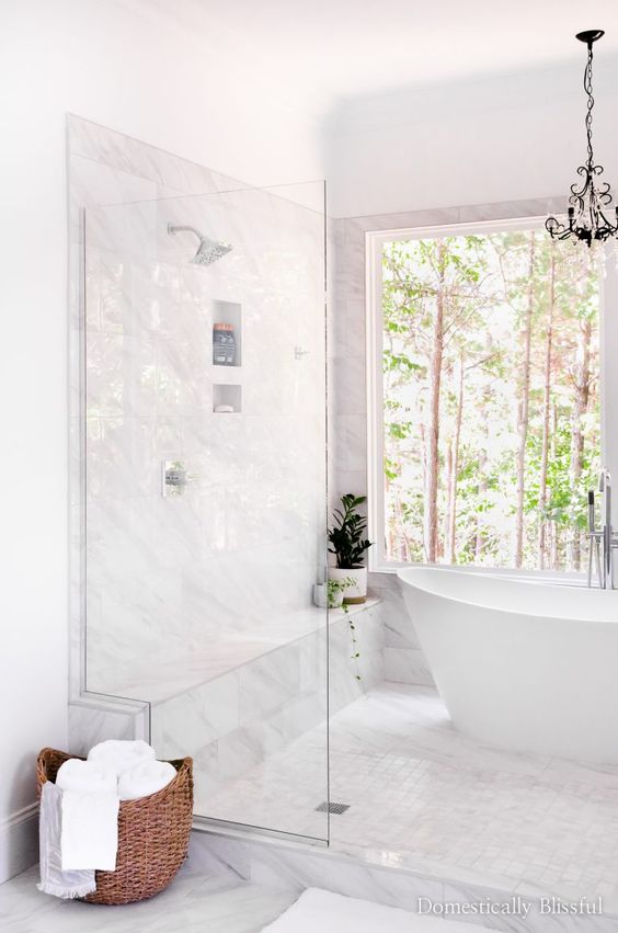 a beautiful white bathroom with a glazed wall and a tub, white marble and Zellige tiles and a chic chandelier
