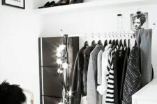 a black and white makeshift closet with open shelves, a metal clothes rack and a white dresser, some lights and a chair
