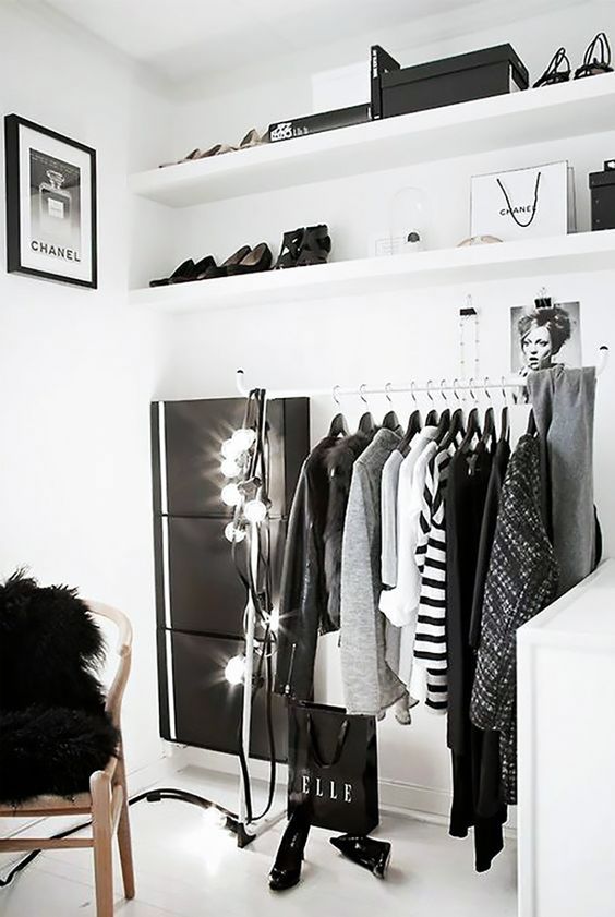 a black and white makeshift closet with open shelves, a metal clothes rack and a white dresser, some lights and a chair