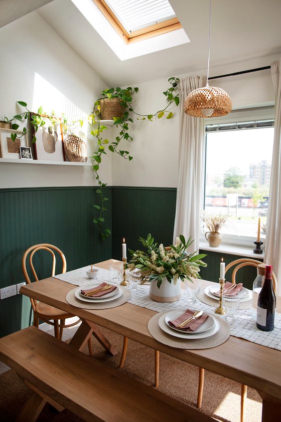 a boho dining room with green beadboard on the walls, a stained table and benches, a woven pendant lamp and greenery