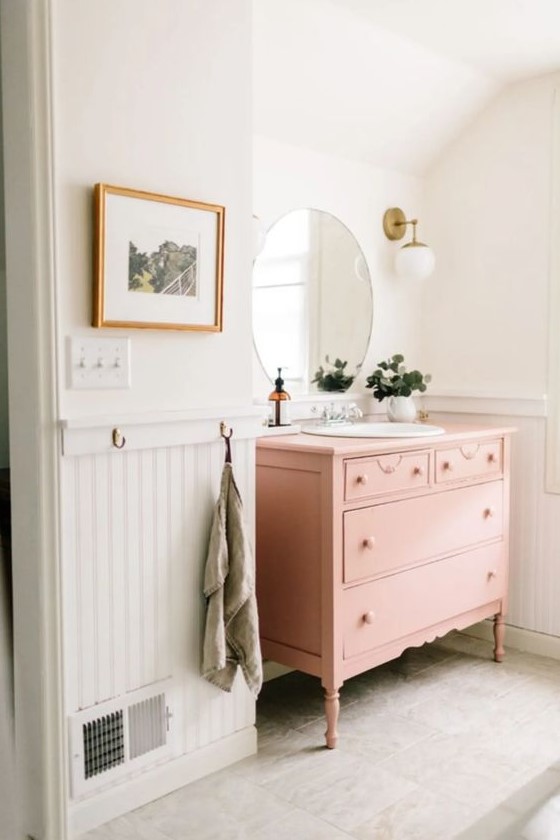 a chic and cute bathroom with white beadboard, a pink vanity, a round mirror, potted greenery and sconces is ethereal