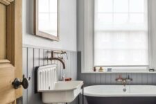a chic bathroom inspried by vintage, with grey beadboard, a black tub, a vintage sink and a large window with shades