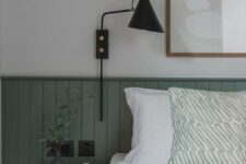 a chic bedroom with green beadboard, a black nightstand and sconce, a neutral artwork and some accessories