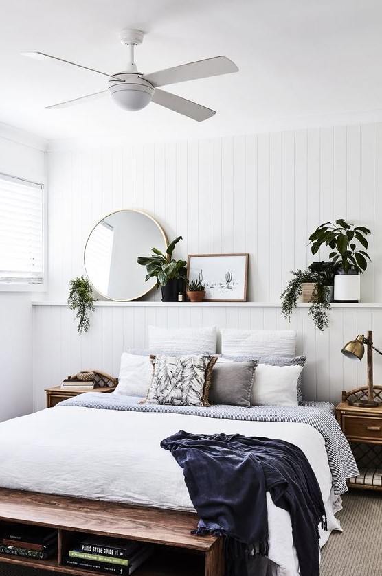 a chic coastal bedroom with a white beadboard wall, wooden furniture, potted plants and greenery, a round mirror and comfy textiles