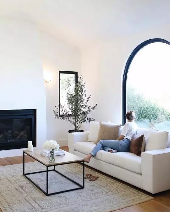 a chic neutral living room with an arched window, a built in fireplace, a white sofa and a coffee table and a potted mini tree