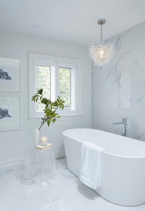 a chic white bathroom clad with marble, an oval tub, a gallery wall, an acrylic table and a bubble chandelier
