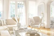 a chic white living room with a sectional, a duo of stone slab coffee tables, a white chair, white textiles and a chic chandelier