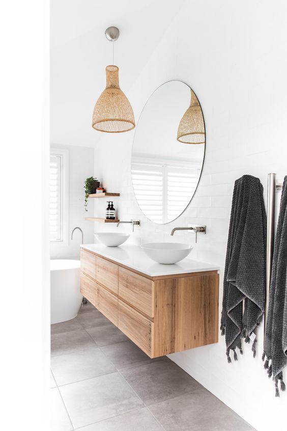 a contemporary bathroom with grey tiles on the floor and white walls, a tub, a floating timber vanity, a round mirror and a woven pendant lamp