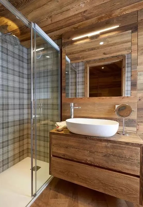 a cool chalet bathroom clad with wood, with a floating vanity, checked tiles in the shower space and a lit up mirror