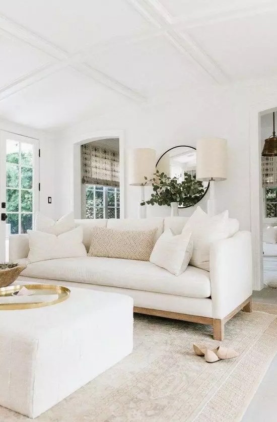a cool living room with a white sofa and a matching ottoman, with a round mirror, two lamps and some chic accessories