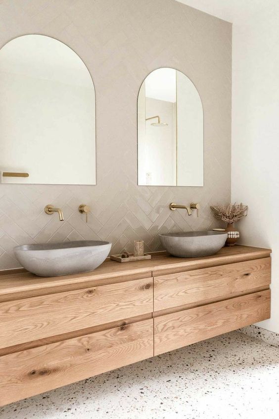 a cool nook clad with glossy grey tiles, arched mirrors, a floating timber vanity, concrete sinks and brass fixtures
