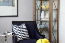 a cool nook with a rough wood storage unit, a navy chair, a yellow ottoman, a printed pillow and a side table