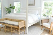 a cozy white bedroom with a frame bed with white bedding, a woven bench and a chair, white nightstands and a rug