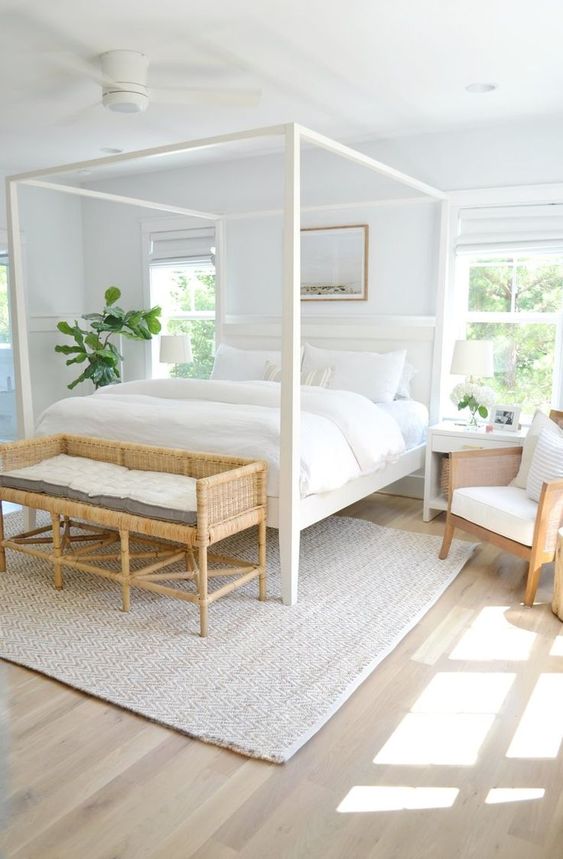 a cozy white bedroom with a frame bed with white bedding, a woven bench and a chair, white nightstands and a rug
