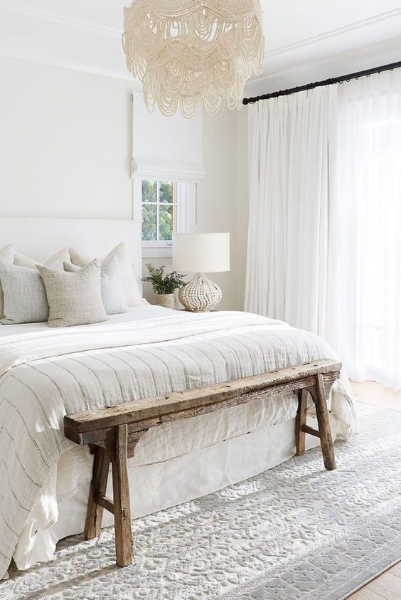 a cozy white bedroom with a white upholstered bed, neutral bedding, creamy curtains and a printed rug, a cool chandelier