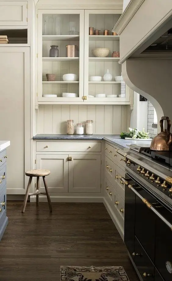 a creamy kitchen with shaker and glass cabinets, a matching beadboard backsplash, grey stone countertops and a large hood