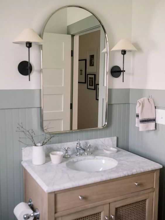 a delicate farmhouse bathroom with grey beadboard on the walls, a stained vanity, a mirror and sconces is chic
