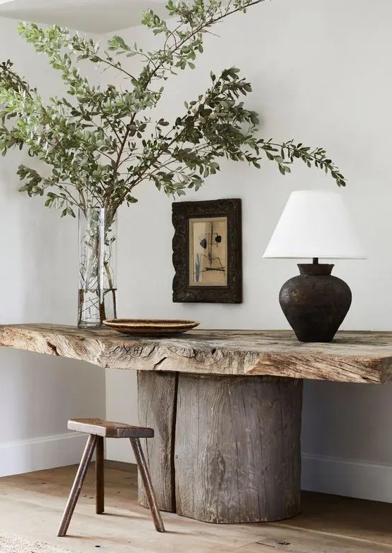 a fabulous rough wood table with a wooden stool, some greenery branches, a black table lamp and an artwork