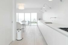 a large and super long minimalist white kitchen with only floor cabinets and whitewashed floors plus lights over the cabinetry