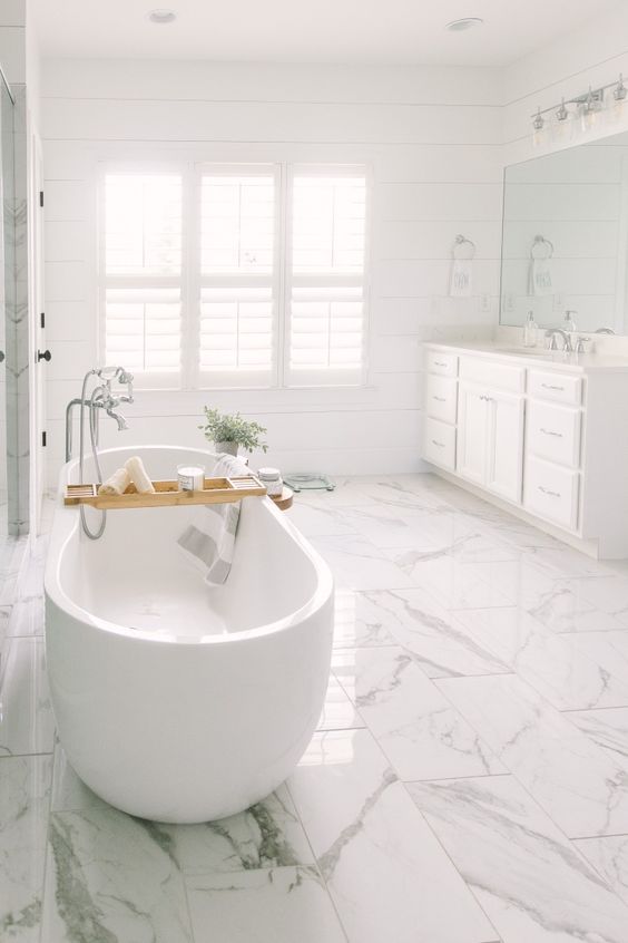 a light filled white bathroom with a large vanity by the window, a white marble tile floor and an oval tub plus potted greenery