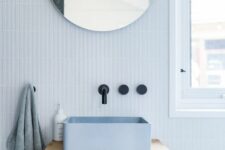 a lovely light blue bathroom with a small floating vanity, a blue square sink, a round mirror, black fixtures and lots of natural light