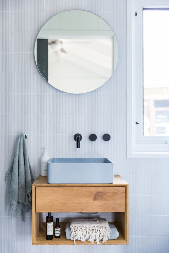 a lovely light blue bathroom with a small floating vanity, a blue square sink, a round mirror, black fixtures and lots of natural light