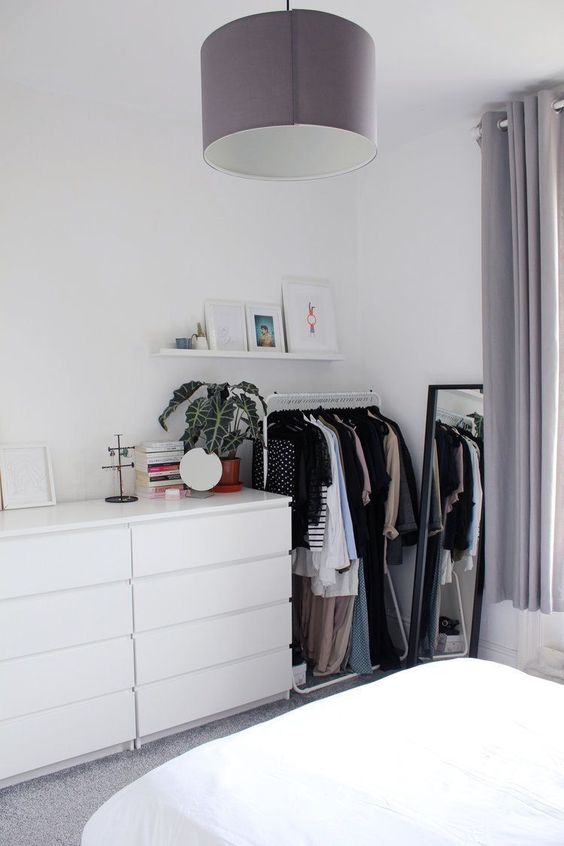 a makeshift closet with a sleek white dresser, a white metal rack for hanging clothes and a floor mirror in a black frame