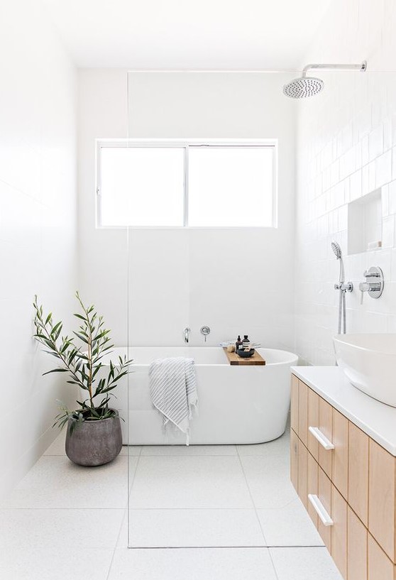 a minimalist airy bathroom with white tiles everywhere, a wooden vanity, a bathtub, a potted plant and a white sink
