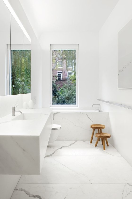 a minimalist white bathroom done with marble, a floating sink and marble clad tub plus a window