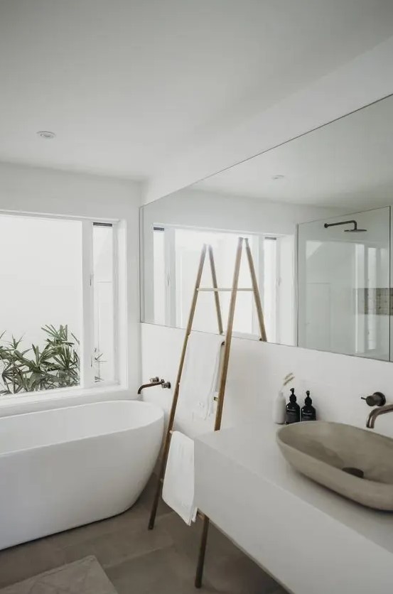 a minimalist white bathroom with a long mirror, a free standing bathtub, a stone sink and a ladder for storage