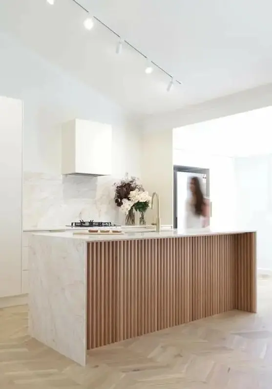 a minimalist white kitchen with a white stone backsplash and countertops and a ribbed kitchen island plus spotlights