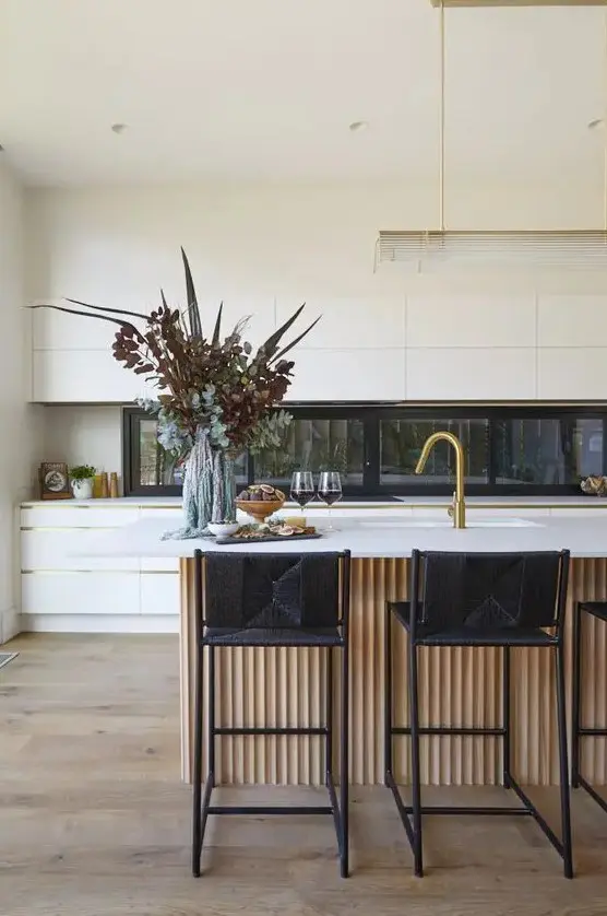 a minimalist white kitchen with a window backsplash, a large fluted kitchen island with black stools and gold touches here and there
