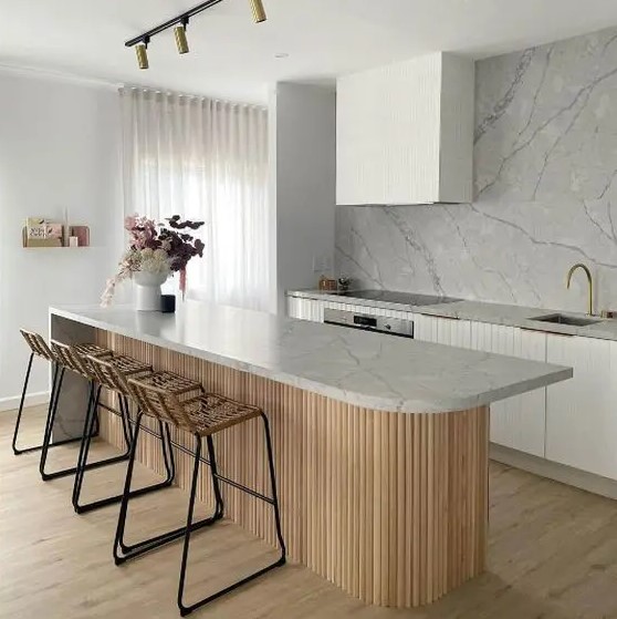 a minimalist white kitchen with fluted cabinets, a white marble backsplash, a curved kitchen island with a stone countertop