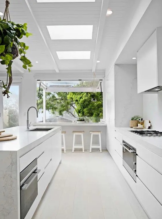 a minimalist white kitchen with marble surfaces and a large window that is extended outdoors as a meal space