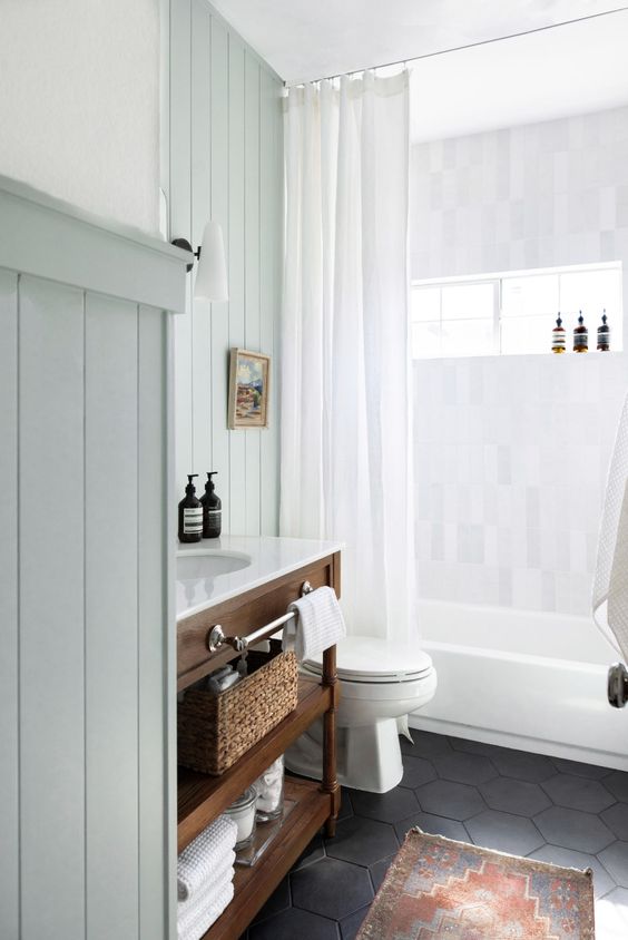 a modern farmhouse bathroom with light green beadboard, a stained vanity, a black hex tile floor and a bold rug