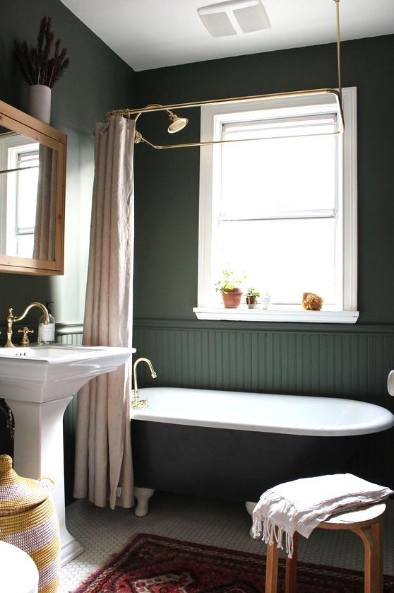 a moody vintage-inspired bathroom with hunter green walls and beadboard, a black tub, a tile floor and a free-standing sink plus touches of brass