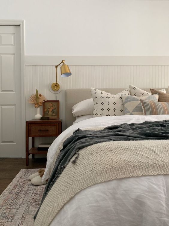 a neutral bedroom with beadboard on the walls, a neutral bed with neutral bedding, a vintage nightstand and a brass sconce
