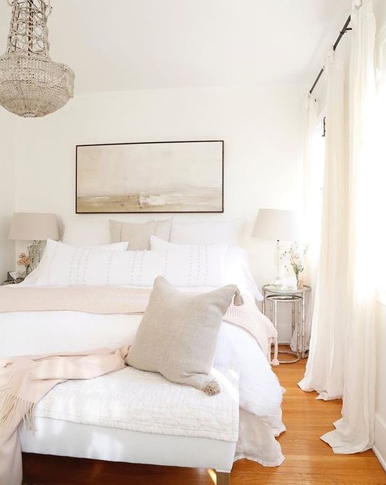 a neutral chic bedroom with a white bed and bench, shiny glass nightstands, lamps and a gorgeous crystal chandelier