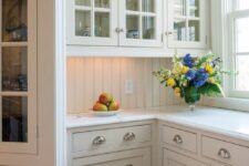 a neutral cottage kitchen with shaker and glass facade cabinets, white stone countertops and a creamy beadboard backsplash