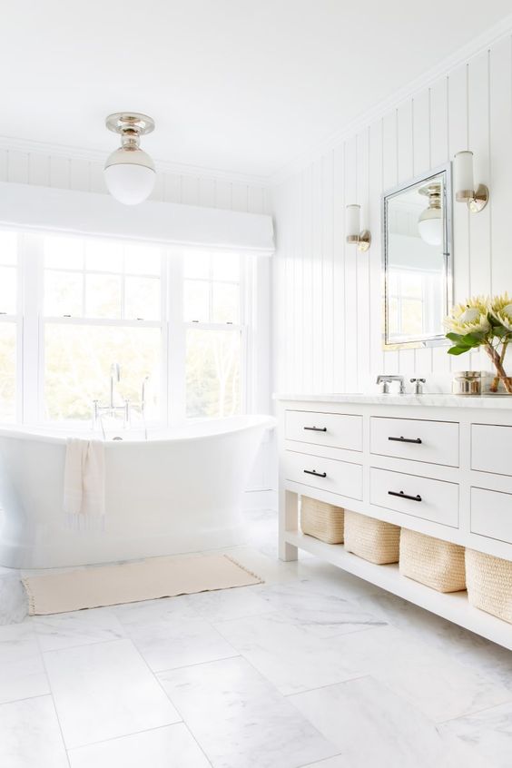 a neutral farmhouse bathroom with a tub by the window, a large vanity, baskets for storage, white marble tiles