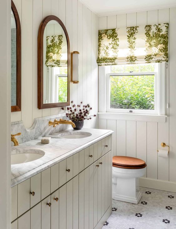 a neutral rustic bathroom clad with beadboard, with a matching double vanity, a couple of arched mirrors and a printed curtain