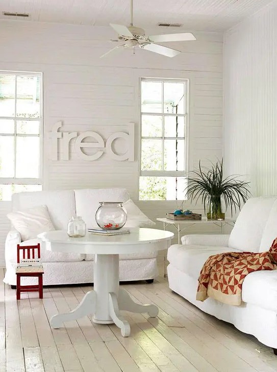 a pretty white living room with white seating furniture, a round table, greenery, a metal coffee table and letters on the wall
