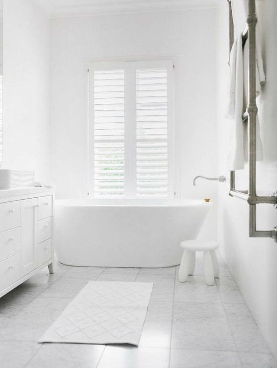 a pure white bathroom with a free-standing tub, a small stool, a large vanity and marble tiles on the floor