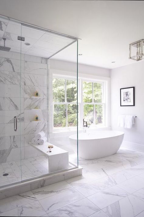 a refined white bathroom fully clad with white marble tiles, with a large shower space and an oval tub by the window