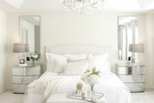 a refined white bedroom with an upholstered bed, white bedding, mirror nightstands, a chic chandelier and taupe lamps