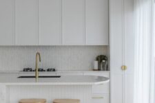 a refined white kitchen with shaker style cabinets, a slatted backsplash and a kitchen island, stained stools and brass handles