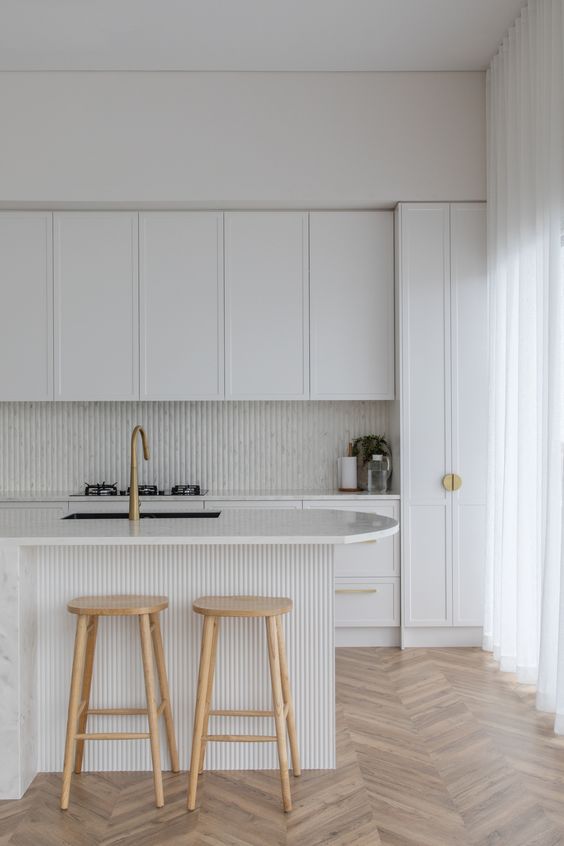 a refined white kitchen with shaker style cabinets, a slatted backsplash and a kitchen island, stained stools and brass handles