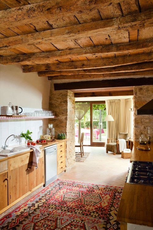 a rustic kitchen with a rough wood ceiling and beams, wooden cabinets and countertops, a printed rug and stone walls