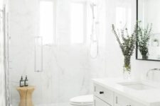 a serene white bathroom with a shaker style vanity, a shower with two windows, a wooden stool in the shower and a mirror